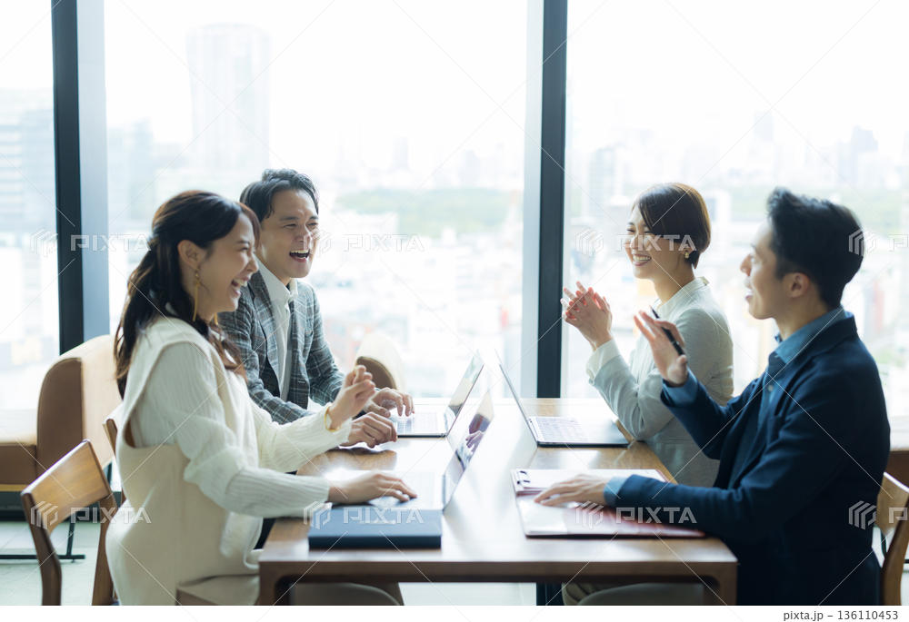 窓際の会議テーブルで4名が打ち合わせをしている様子。ノートPCと資料を広げ、話しながら笑っている。日中の自然光、引きの構図