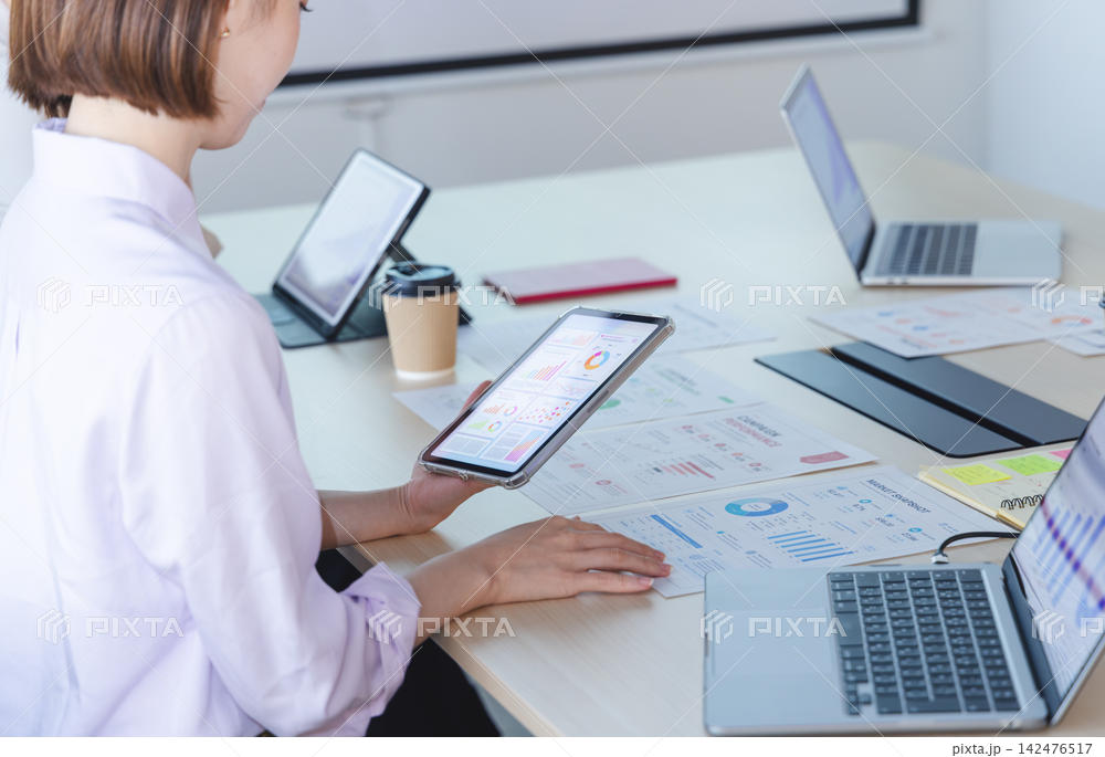 デスクにタブレットとノートPC、印刷したデータ資料を広げて内容を確認する担当者。斜め後方からの俯瞰、室内の自然光